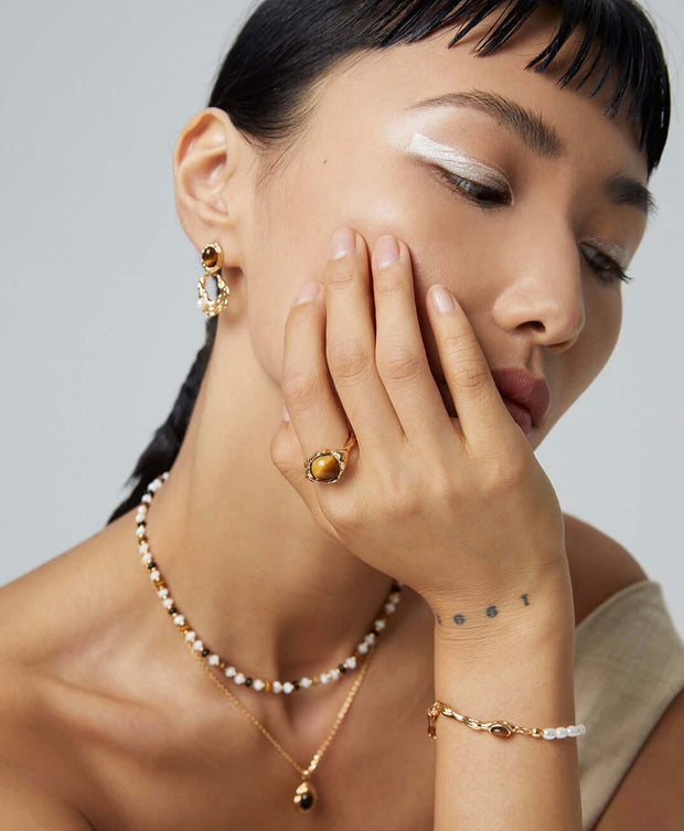 A woman wearing a necklace and earrings with pearls, adorned with a Tiger's Eye Ring of adjustable size.