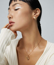 A woman wearing a white sweater and gold earrings with the Phoenix Grace Necklace, which has 18K gold plating.